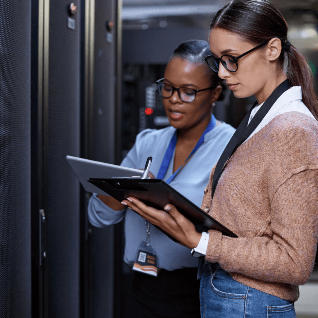 Two women IT professionals holding and looking at clipboards in front of large computers
